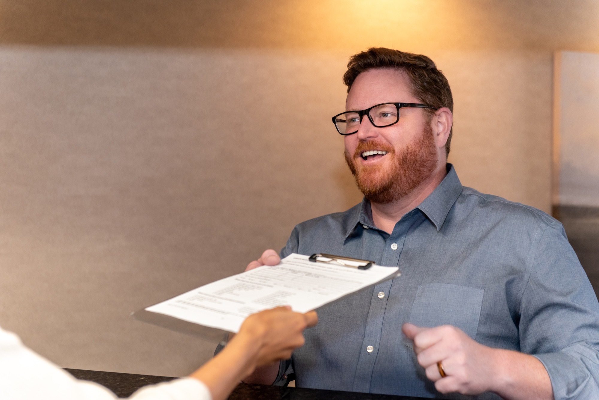A patient at the front desk taking a new patient form on a clipboard handed to him across the counter