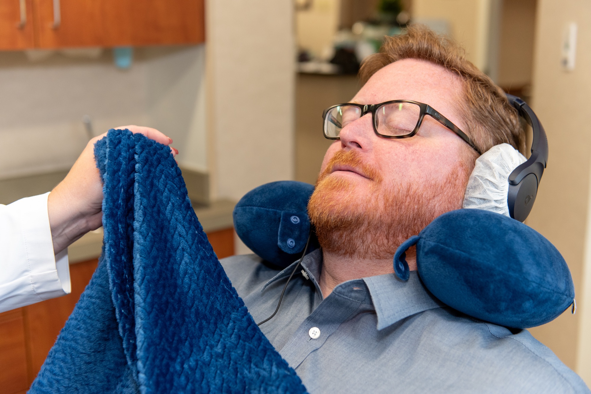 A patient resting in the chair with a neck pillow and noise-cancelling headphones while Dr. Lokitis lays a soft blanket over him