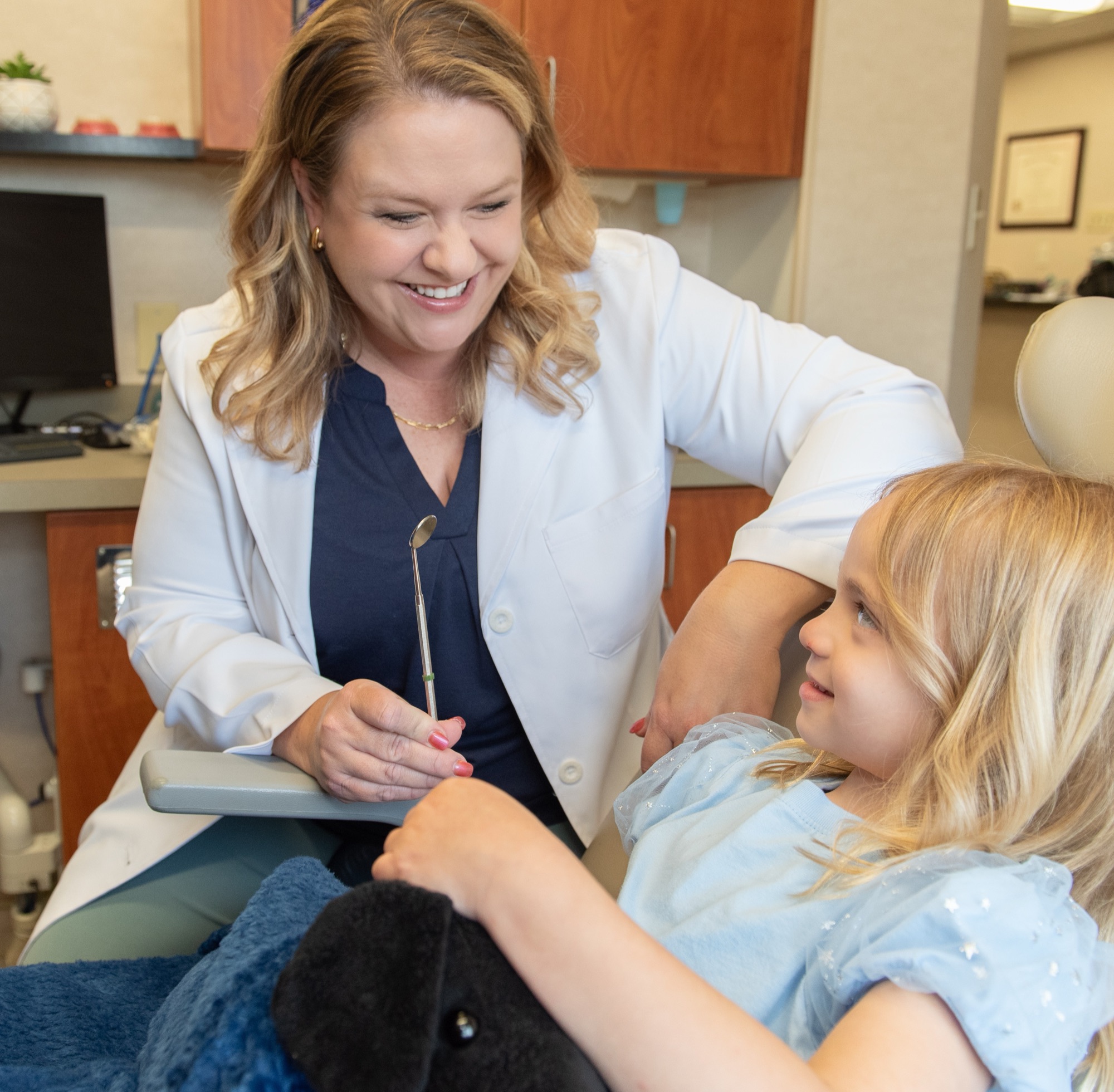 Dr. Julia Lokitis leaning in to talk with a young girl in the dental chair, a soft blanket over her lap